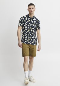 Camisa de manga corta abotonada en color crema con un patrón floral en azul marino y amarillo, combinada con pantalones cortos color caqui. Calcetines blancos y zapatillas de color claro.