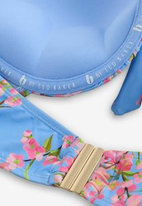 Nahaufnahme eines blauen Bikini-Oberteils mit pinken Blumen, goldenem Verschluss und der Aufschrift "b by Ted Baker" an der inneren Naht.