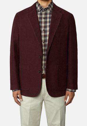 Mężczyzna w ciemnym bordowym marynarce, wielokolorowej koszuli w kratę zapinanej na guziki, beżowych spodniach i brązowym pasku.