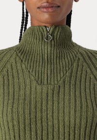 Suéter de punto acanalado verde con cuello alto y una cremallera corta con un tirador circular. Tejido texturizado con líneas verticales.