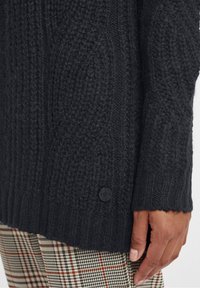 Mörkgrå stickad tröja med ett mönstrat textur, försedda med en svart knapp på vänster ärm, i kombination med rutiga byxor.