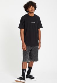 Svart bomull t-skjorte med "VOLCOM" logo, parret med grå denimshorts, svarte sneakers og svarte sokker med logoer. Enkelt, uformelt antrekk.