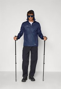 Navy waterproof jacket with hood, zip closure, two front pockets; paired with black pants and trekking poles. Worn with sunglasses.