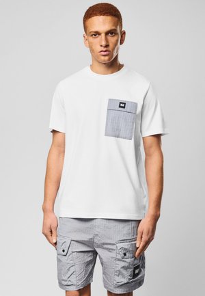 Mężczyzna w białej koszulce z szarą kieszonką i pasujących szarych szortach cargo, stojący na jednolitym jasnoszarym tle.