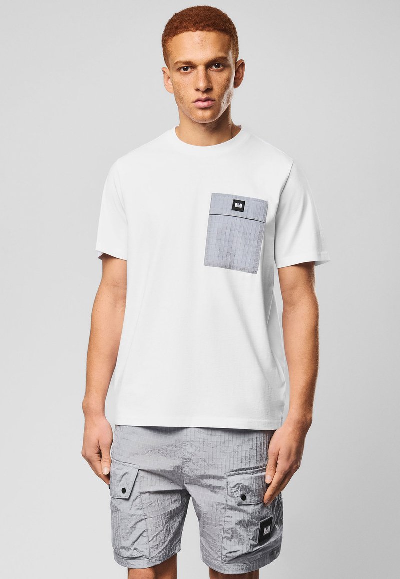Homme portant un T-shirt blanc avec une poche grise et un short cargo gris assorti, debout devant un fond gris clair uni.