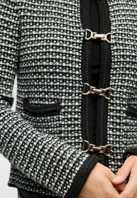 Schwarz-weißes Tweed-Jacket mit strukturiertem Gewebe, ausgestattet mit goldfarbenen Verschlüssen und zwei Taschen vorne.