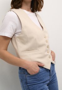 Gilet beige con design sartoriale, chiusura a cinque bottoni e una tasca laterale, indossato sopra una camicia bianca con jeans blu. Tessuto dalla consistenza liscia.