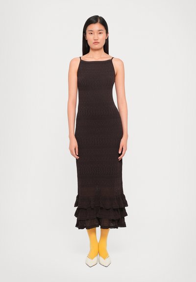 Dunkelbraues Strickkleid mit einem taillierten Oberteil und gestuften Rüschensaum. Getragen mit gelben Socken und weißen, spitzen Schuhen. Schlichte Träger.