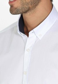 Chemise blanche à boutons avec un col marine contrastant. Présente un design de col pointu et une texture subtile. Les boutons sont ronds et transparents.