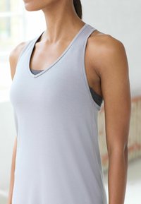 Szary sportowy top bez rękawów z dekoltem w kształcie litery V i cienkimi ramiączkami. Charakteryzuje się gładką teksturą oraz warstwowym wyglądem z ciemnym podkładem.
