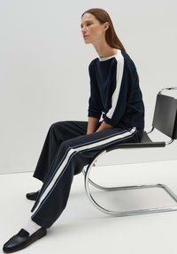 Ensemble de sport marin avec un haut à manches trois-quarts doté de rayures blanches et un pantalon large assorti. Chaussures noires à enfiler et chaise en chrome.