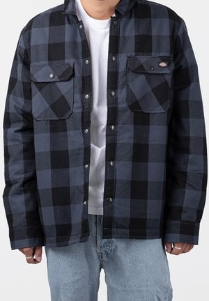 Mann trägt blau-schwarz kariertes Hemd mit Knöpfen über weißem T-Shirt und hellblauen Jeans, Hände locker an den Seiten.