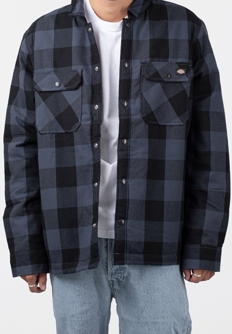 Homme portant une chemise à carreaux bleue et noire boutonnée sur un t-shirt blanc avec un jean bleu clair, les mains détendues de chaque côté.