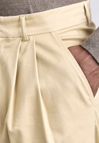 Beige bukser laget av mykt stoff har en plisserte design, en linning med sløyfe og sidelommer. Overflaten ser jevn ut.