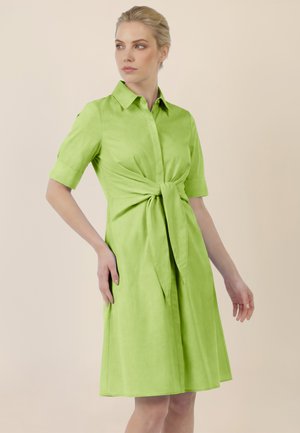 Frau trägt ein knielanges hellgrünes Kleid mit kurzen Ärmeln, Kragen und einer gebundenen Schleife in der Taille, steht vor einem einfarbigen beigen Hintergrund.