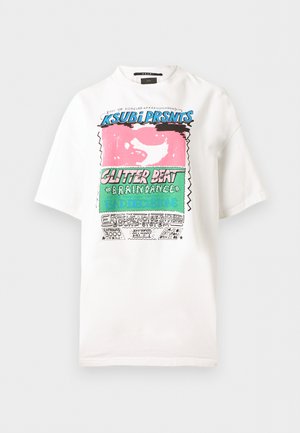 Kobieta o długich ciemnych włosach, ubrana w białą koszulkę z grafiką, czarny pasek, ciemne jeansy z wysokim stanem i czarne buty na obcasie z odkrytymi palcami, stojąca z rękami za plecami.