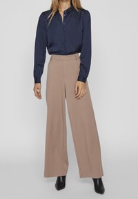 Camicia in satin blu navy con bottoni e maniche lunghe, abbinata a pantaloni beige a gamba larga e stivali neri alla caviglia. Tessuto liscio con vestibilità su misura.