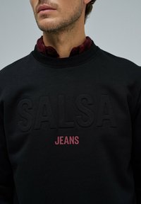 Sudadera negra con estampado en relieve de "SALSA" y "JEANS" en letras rosas. Cuello acanalado y textura indicativa de un material suave.