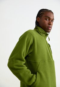 Grønn fleece-pullover med en kvart glidelås krage, som har en teksturert overflate og en frontlomme. Designet inkluderer lange ermer og en avslappet passform.