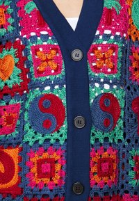Cardigan all'uncinetto con vivaci quadrati in rosso, rosa, verde e blu. Bordo a coste blu navy con due bottoni neri rotondi sul frontale.