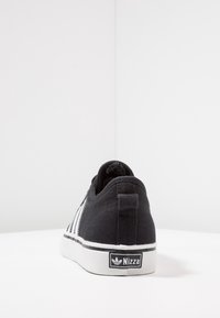 Czarne niskie sneakersy z białą podeszwą, trzy białe paski po boku i logo "Nizza" z tyłu, pokazane z tylnego kąta.