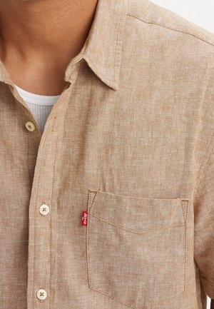 Beige linen button-up shirt with front pocket and red Levi’s tag worn over a white ribbed undershirt on a person’s upper torso.