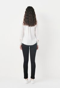 Blusa blanca de manga larga con un dobladillo curvado, combinada con pantalones ajustados negros y zapatos blancos con detalles metálicos.