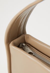 Beżowa skórzana torebka o gładkiej fakturze, z metalowym suwakem i detalem z logo. Projekt zawiera usztywnione otwarcie.