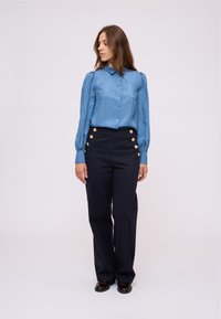 Blusa di seta azzurra chiara con chiusura a bottoni, caratterizzata da maniche a palloncino. Pantaloni alti di colore blu scuro con bottoni dorati sui lati.