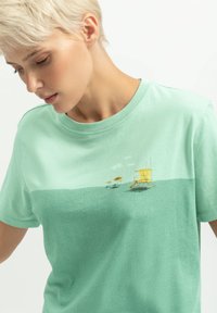 Mintgroene t-shirt met een grafische print van gele lifeguardtorens en parasols. Gemaakt van zacht, glad katoenmateriaal. Korte mouwen.