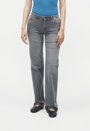 Persoon die grijze rechttoe-rechtaan jeans, een blauw topje en zwarte platte schoenen draagt, staand tegen een egale witte achtergrond.