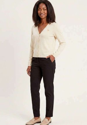 Femme debout, la main dans la poche, portant un cardigan crème, un pantalon noir et des mocassins beiges, souriant à la caméra sur un fond uni.