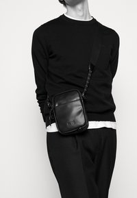 Person, der einen schwarzen Pullover, schwarze Hose und eine schwarze Leder-Umhängetasche mit dem "HUGO"-Logo trägt, vor einem schlichten weißen Hintergrund steht.