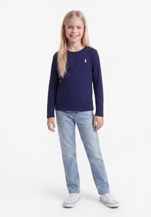Ung pige med langt blondt hår iført en marineblå langærmet trøje, lyseblå jeans og hvide sneakers, smilende og stående.