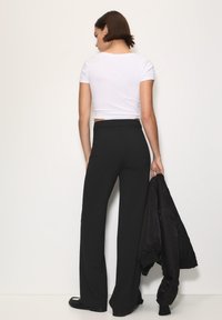 Crne široke hlače sa prugastim uzorkom u kombinaciji s prilagođenim bijelim majicom kratkih rukava, na jednostavnoj pozadini. Crna jakna se drži u ruci.