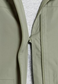 Vaaleanvihreän takin etuosassa on kaksinkertaiset vetoketjut ja nepparikiinnitys vaaleanharmaan paidan päällä.