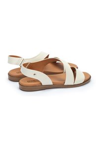 Sandalias de cuero blanco con diseño de tiras cruzadas, talón abierto y suela texturizada marrón. Presenta un pequeño acento metálico cerca de la correa del talón.