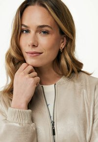 Jasno beżowa kurtka bomberka z prążkowanymi mankietami i zamkiem z przodu. Gładka, lekko błyszcząca tekstura. Modelka dotykająca brody, nosząca proste kolczyki.