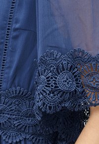 Σκούρο μπλε ύφασμα με περίτεχνη δαντέλα με λουλουδένιο μοτίβο σε διαφανές μανίκι, που δείχνει λεπτές υφασμένες ραφές και διακόσμηση.