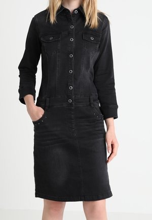 Czarna sukienka z denimu zapinana na guziki, z długimi rękawami, kieszeniami na piersiach i rękami w bocznych kieszeniach, noszona przez stojącą osobę.