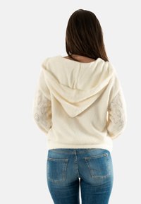 Felpa crema con cappuccio testurizzato e maniche a maglia con motivo, abbinata a jeans blu aderenti con tasche posteriori. Tessuto morbido e accogliente.