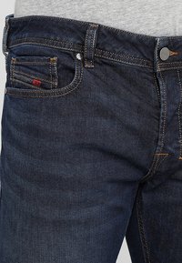 Jeans de mezclilla azul oscuro con un ajuste slim. Incluye un bolsillo frontal, costuras naranjas y una etiqueta de marca en el bolsillo trasero.