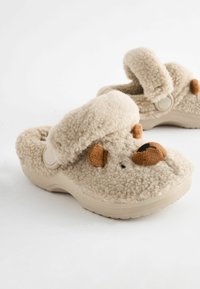 Zapatos tipo zueco de felpa en beige con una textura suave, que cuentan con un puño acolchado y detalles de cara de oso, incluyendo orejas y nariz marrones.