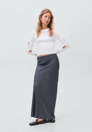 Top bianco a maniche lunghe con dettagli a taglio abbinato a una gonna lunga a scacchi blu e nera. La gonna presenta una texture liscia. Scarpe nere senza tacco.