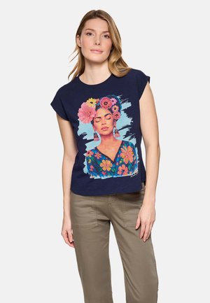 Mujer con cabello castaño claro que lleva una camiseta azul marino con un retrato colorido de una mujer con los ojos cerrados y una corona de flores, combinada con pantalones beige.