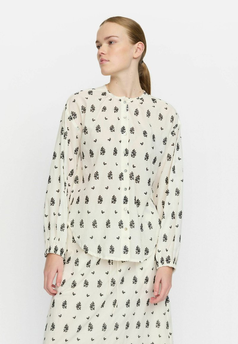 Camicia leggera a fantasia floreale in crema con disegni neri, maniche lunghe, orlo arrotondato e chiusura con bottoni. Tessuto morbido e semitrasparente.