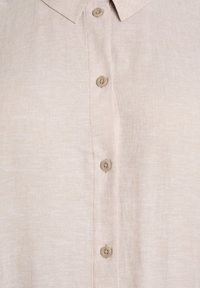 Beige pellavapaita, jossa on neljä vaaleanruskeaa pyöreää nappia ja klassinen kaulus, joka näyttää kankaan rakenteen ja ompelun yksityiskohdat.