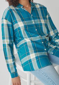 Camicia a quadri turchese con davanti a bottoni, caratterizzata da accenti bianchi e gialli. Tessuto morbido, vestibilità rilassata e maniche lunghe.