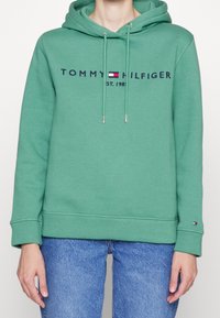 Osoba v zelené mikině Tommy Hilfiger s logem a vlajkou na hrudi a rukávu, v kombinaci s modrými džíny na bílém pozadí.