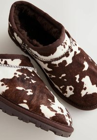 Papuci cu imprimeu de vacă maro și alb, cu un exterior moale din blană, design slip-on și tălpi din cauciuc pentru aderență.
